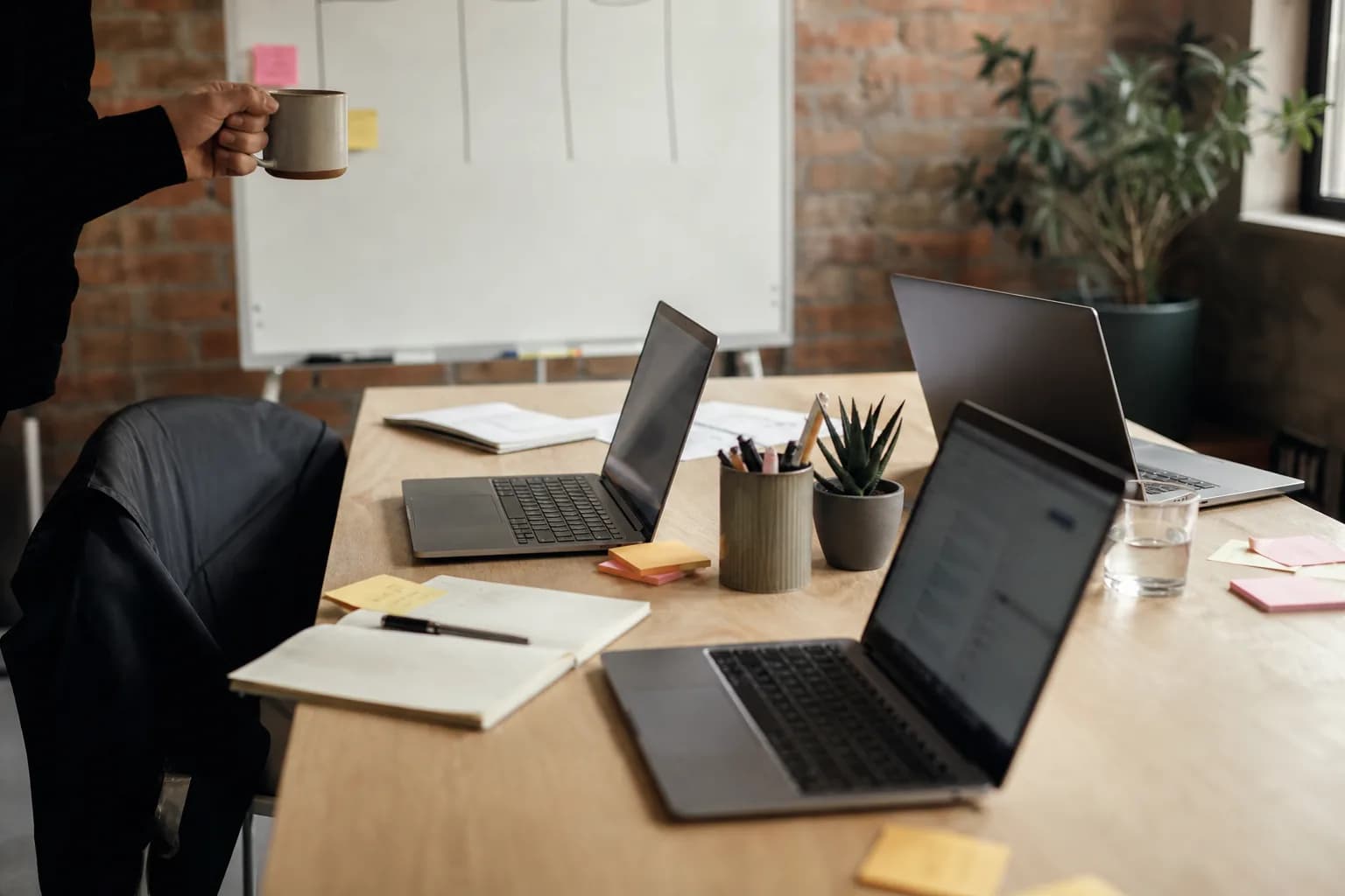 Small agency workspace with a long shared table, three laptops with blurred screens, sticky notes, a whiteboard with empty columns, one off-frame hand holding a mug, afternoon light, brick wall, and no faces.